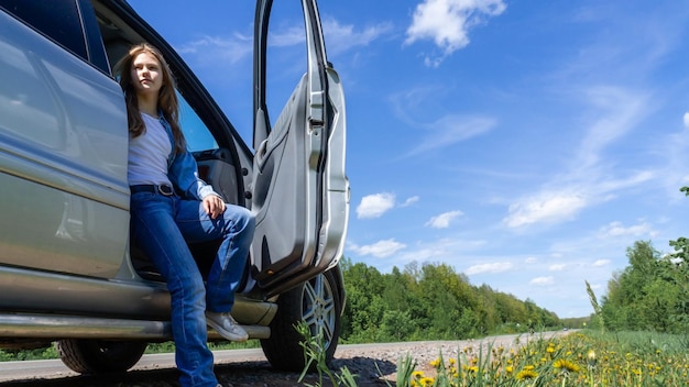 A young girl with blonde hair is sitting in a car with the door open on the side of the road outside