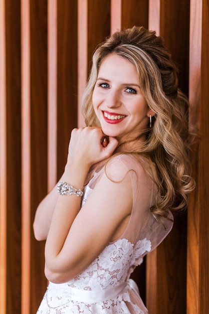 Young girl with blonde long hair in white wedding dress is standing near wood wall in hotel lobby Fashion happy bride vertycal photo