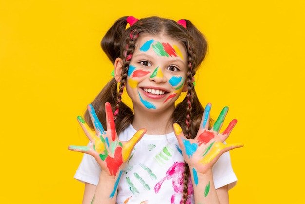 A young girl with brightly painted hands and multi colored pigtails shows her palms smiling broadly yellow isolated background