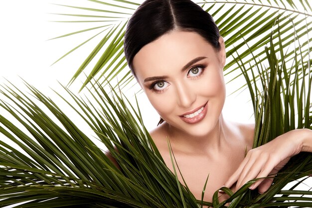 Foto giovane ragazza con capelli castani fissati dietro, pelle pulita e fresca, grandi occhi e spalle nude in posa con foglie verdi al muro bianco, ritratto, sorridente.