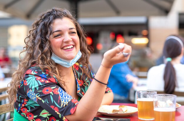 Young girl with curly hair who laughs while sitting drinking beer in a bar, wearing a protective mask under her chin, concept of resuming social activities, optimism