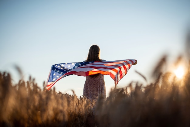 写真 バックグラウンドで夕日を背景にライ麦畑でアメリカの国旗を持つ少女。アメリカの独立記念日のコンセプト