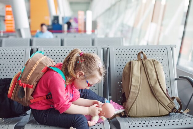 Young girl with glasses playing with a doll at the airport, traveler