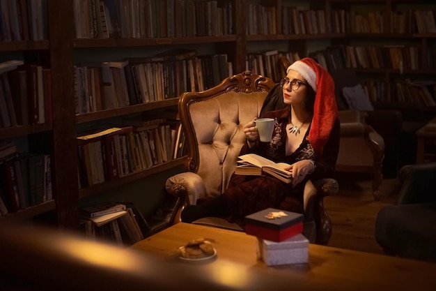 Young girl with glasses in Santa hat drinking cofee while reading a book in a library