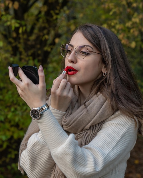 a young girl with glasses stands on a white bridge and paints her lips with red lipstick