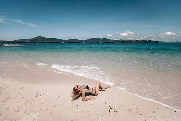 Young girl with a gorgeous body is resting on the beach with white sand near the ocean. Beautiful sexy model in a black bathing suit  and black sunglasses sunbathing.