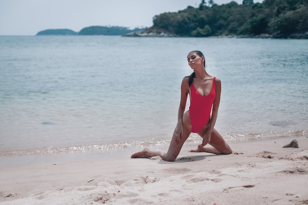 Young girl with a gorgeous body is resting on the beach with white sand near the ocean. Beautiful sexy model in a red bathing suit sunbathing. Brunette with curly hair in bikini.