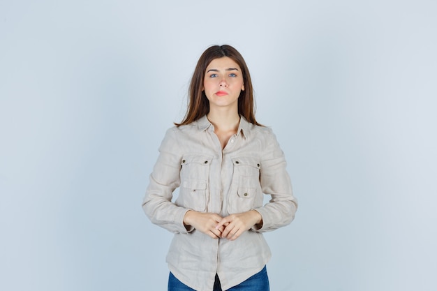 Young girl with hands in front of her, biting lips in beige shirt, jeans and looking worried , front view.