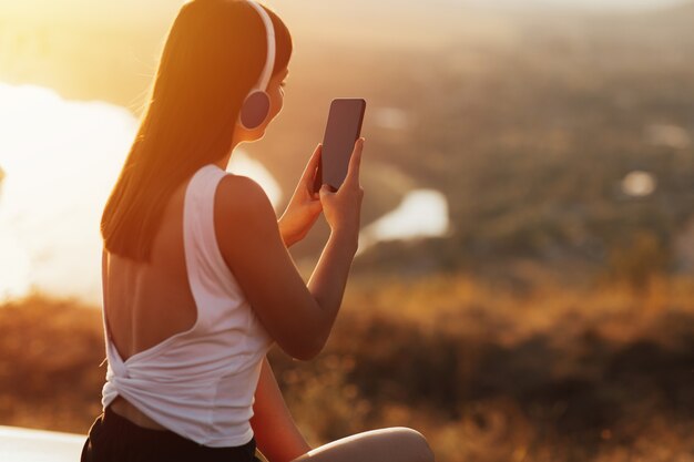 Young girl with headphones listening to music from her smart phone on the hill during at sunset.