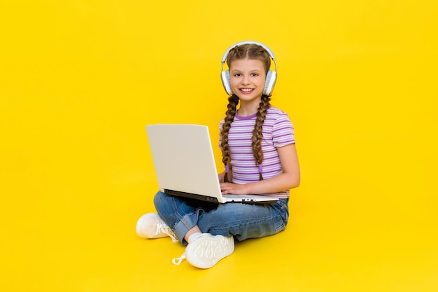 A young girl with a laptop a little girl is sitting crosslegged on the floor with headphones on and holding a laptop in her hands social networks for teenagers yellow isolated background