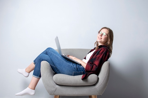 Молодая девушка с ноутбуком сидит на мягком удобном кресле и улыбается, женщина пользуется компьютером у белой глухой стены, она фрилансером и печатает текст, копирует пространство