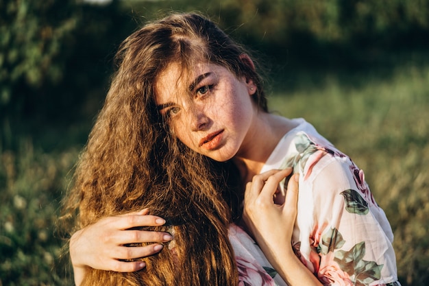 Young girl with long curly hair, brunette, face in freckles, with makeup and green eyes, in light white dress posing