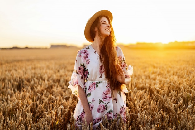 Молодая девушка с длинными вьющимися волосами и веснушками на лице, в шляпе, в светлом белом платье с цветочным принтом, стоя на пшеничном поле, позирует