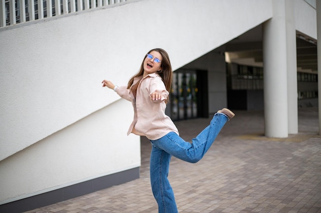 メガネとジーンズを着た長い髪の若い女の子が街の空中で嬉しそうに飛び跳ねる