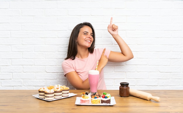 Молодая девушка с большим количеством различных мини-пирожных, указывая указательным пальцем отличная идея