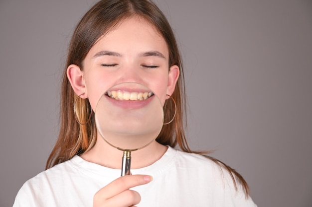 Young girl with magnifying glass smiling excited over gray background Teengirl showing her teeth