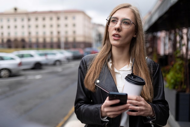 Молодая девушка с мобильным телефоном в руках и стаканом кофе ищет дорогу в городе