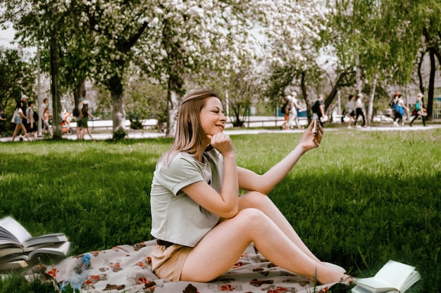 A young girl with a mobile phone in the park takes a selfie photo or watches a video on a modern gadget with WiFi in outdoor public places during a break at the university