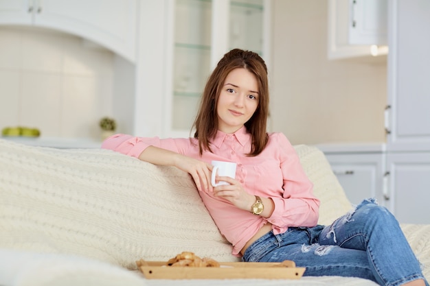 温かい飲み物のマグカップを持つ若い女の子が明るい部屋に座っています。