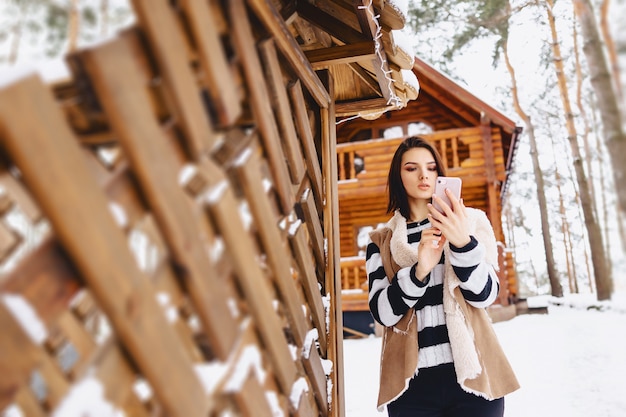 冬の森の木造コテージに対してのチョッキで携帯電話を持つ少女