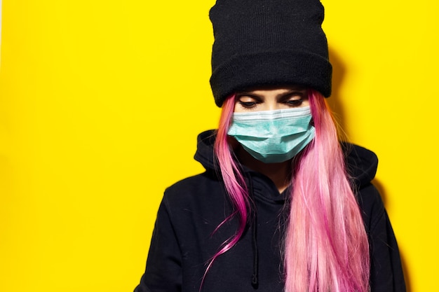 Young girl with pink hair and blue eyes, wearing medical flu mask, dressed in black hoodie sweater and beanie hat on yellow wall.