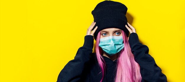 ピンクの髪と青い目をした少女は、医療用インフルエンザマスクを着用し、黄色の壁に黒いパーカーのセーターとビーニー帽子をかぶっています。