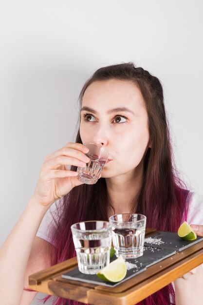 Young girl with pink hair drinks shot of tequila and holds in hand wooden tray with limes, salt and shots on grey background