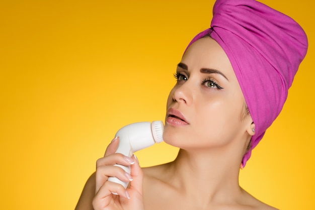 A young girl with a pink towel on her head makes a deep cleansing of her face with an electric brush