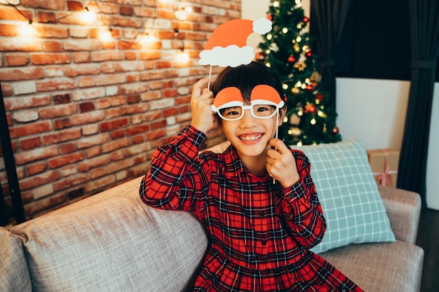 young girl with red dress making funny face playing with fake paper decoration looking at camera smiling attractive. cute kid sitting on sofa at home with christmas tree in background peaceful night.