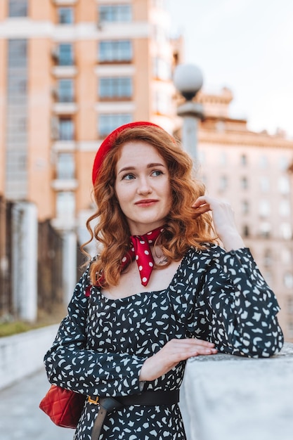 A young girl with red hair in a black dress and red beret against the background of the city