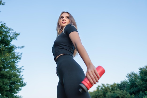 Una giovane ragazza con una figura sportiva distoglie lo sguardo tenendo una bottiglia d'acqua nelle sue mani contro il cielo