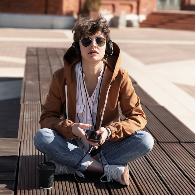 A young girl with a stylish short haircut sits on a bench and listens to music on headphones.
