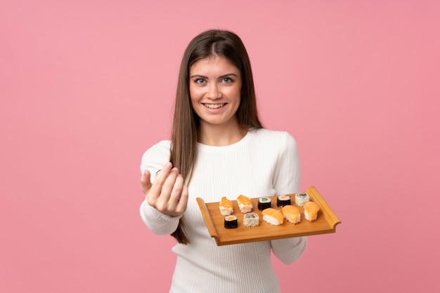 Young girl with sushi over isolated pink  inviting to come with hand. Happy that you came