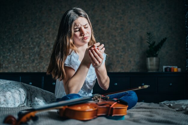 A young girl with a violin sits on the bed and holds a wounded finger with her hand