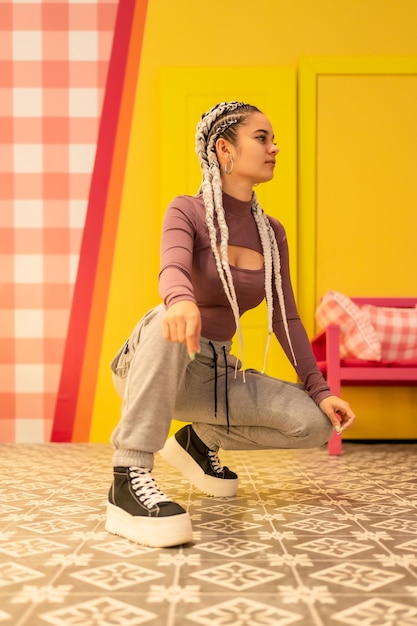 Photo young girl with white braids on a yellow background, crouching in an alternative girl pose or trap dancer or rapper, looking to the sides