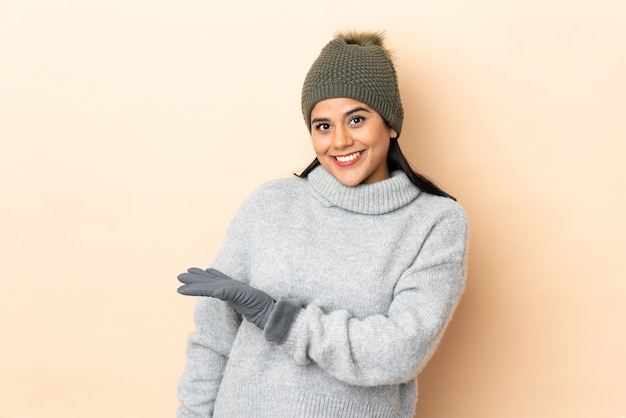 Young girl with winter hat on beige wall extending hands to the side for inviting to come