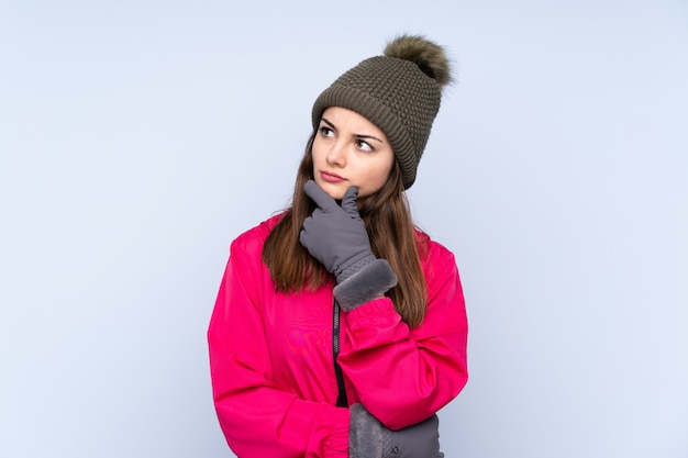 Young girl with winter hat isolated on blue having doubts and with confuse face expression