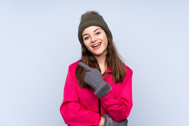 Young girl with winter hat isolated on blue wall surprised and pointing side