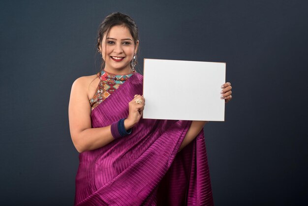 A young girl or woman wearing an Indian traditional saree holding a signboard in her hands on gray background