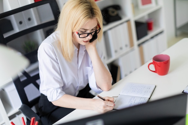 A young girl working at a computer in the office and talking on the phone.