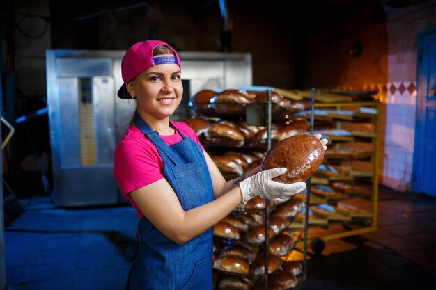 Молодая девушка работает в пекарне. Ставит хлеб на полку. Пекарь женщины на рабочем месте в пекарне. Профессиональный пекарь держит в руках хлеб. Концепция производства хлеба