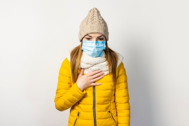 Young girl in a yellow jacket and hat with a medical mask on her face holding her hand to her chest and coughing