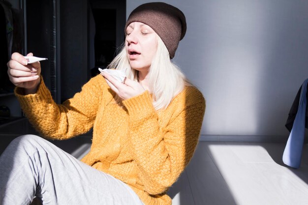 A young girl in a yellow sweater and a brown hat holds a thermometer in her hand and sneezes