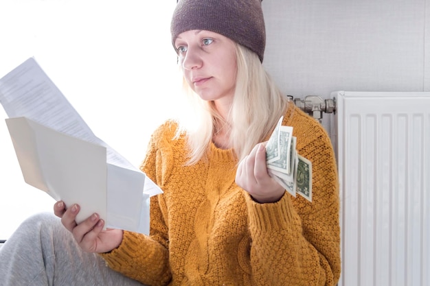 A young girl in a yellow sweater and a brown hat is sitting on the floor counting money and thinking how to pay bills and taxes near a heater with a thermostat