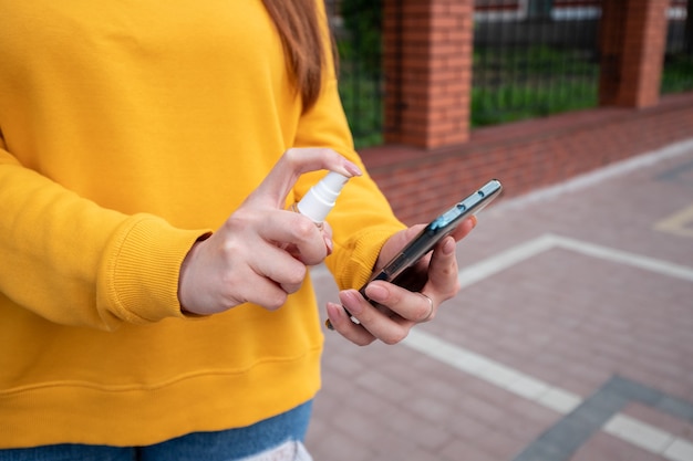 Молодая девушка в желтом свитере дезинфицирует свой телефон дезинфицирующим спреем