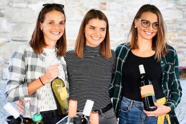 Young girls of family of winemakers selling bottles of wine on street market. Three smiling happy sisters and female friends. Holiday celebration. Leisure together. Summer fun. Farmers at work.