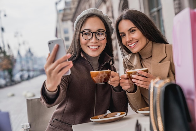 カフェで屋外に座ってコーヒーを飲み、スマートフォンで自分撮りをしている若い女の子の友達