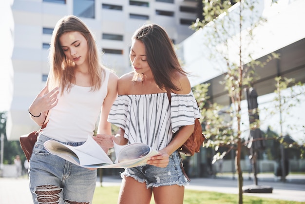 Young girls reading city map and looking for hotel. Lovely tourists with backpacks determine the concept of knowledge of the world