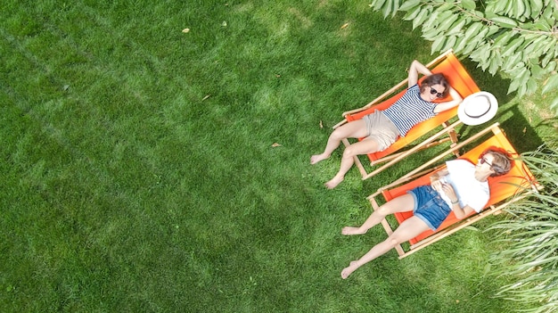 Молодые девушки отдыхают в летнем саду в шезлонгах на траве, подруги веселятся на свежем воздухе