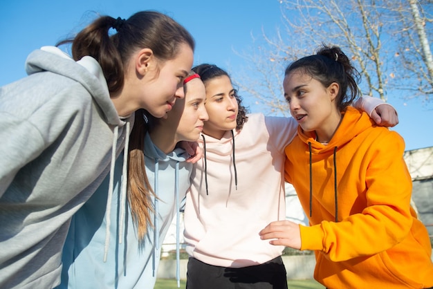 Young girls talking of football game on field. Four happy Caucasian girls in sportswear hugging each other, concentrated on discussing way of winning in game. Team sport and healthy lifestyle concept